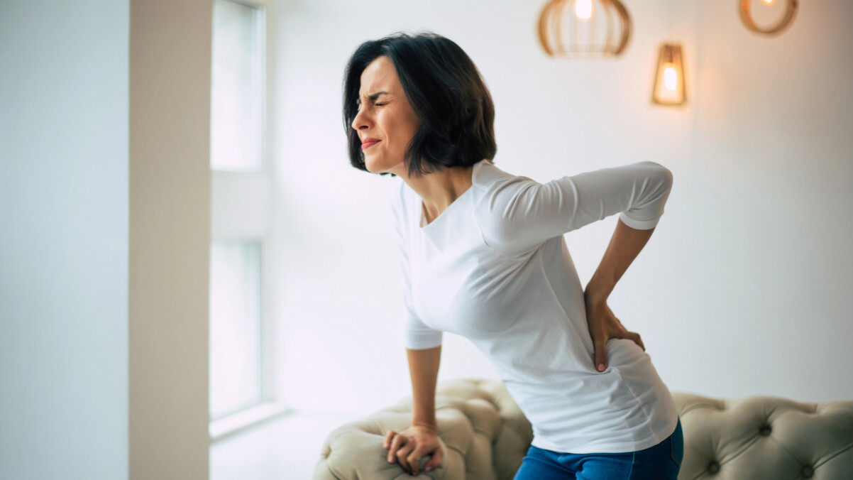 A woman in a white shirt and jeans, experiencing menopause back pain, holds her lower back in discomfort while standing near a sofa in a brightly lit room.