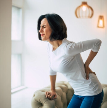 A woman in a white shirt and jeans, experiencing menopause back pain, holds her lower back in discomfort while standing near a sofa in a brightly lit room.