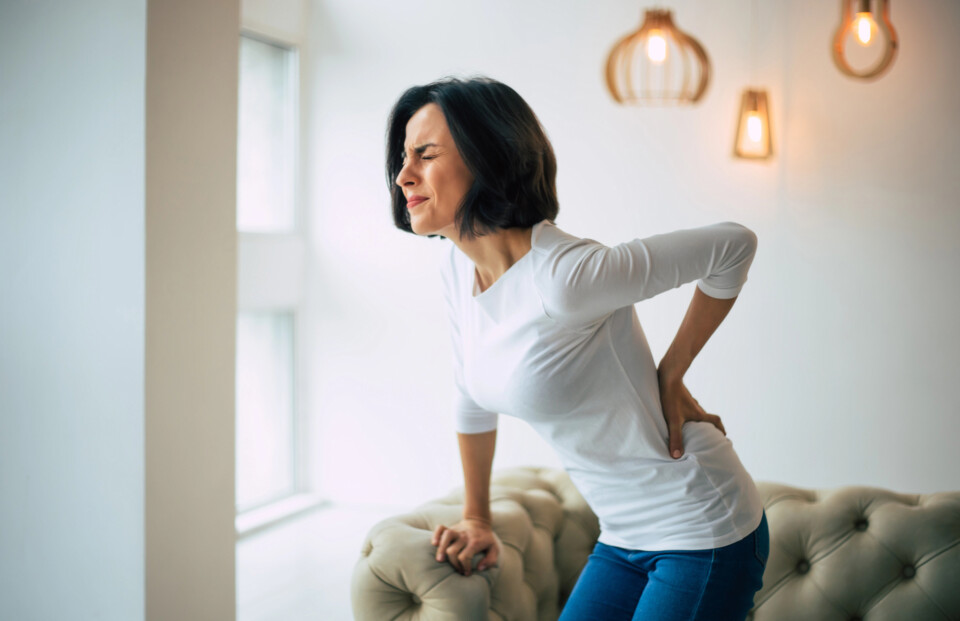 A woman in a white shirt and jeans, experiencing menopause back pain, holds her lower back in discomfort while standing near a sofa in a brightly lit room.