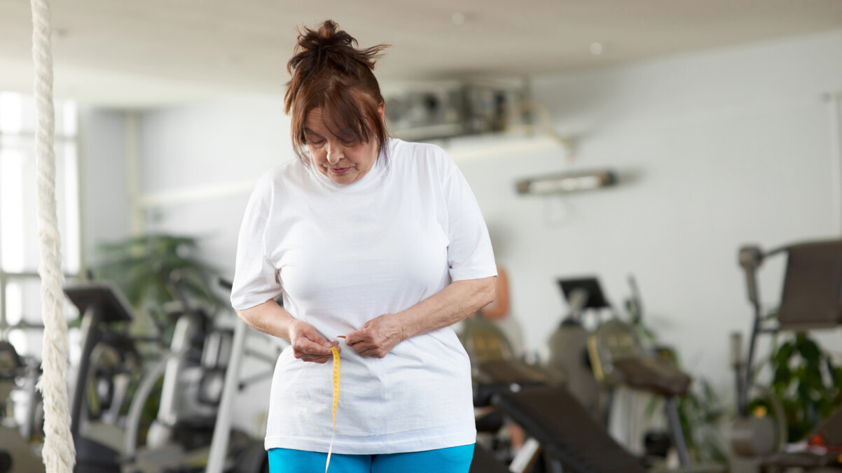 A woman in a gym measures her waist with a tape measure, tracking her weight loss progress amid various exercise equipment, mindful of how menopause can affect fitness goals.