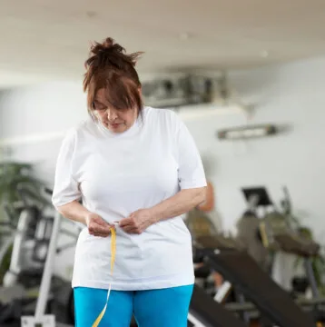 A woman in a gym measures her waist with a tape measure, tracking her weight loss progress amid various exercise equipment, mindful of how menopause can affect fitness goals.