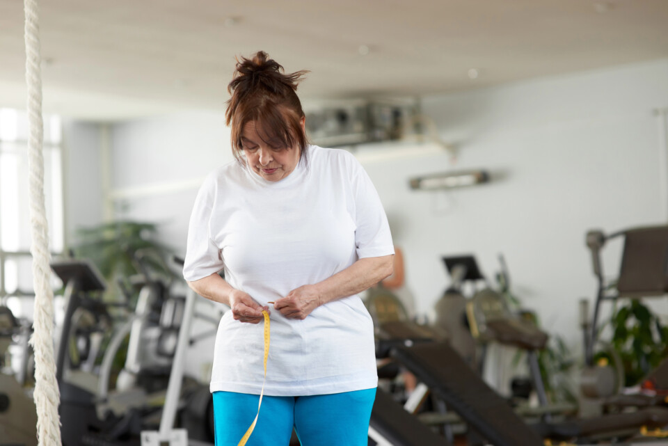 A woman in a gym measures her waist with a tape measure, tracking her weight loss progress amid various exercise equipment, mindful of how menopause can affect fitness goals.