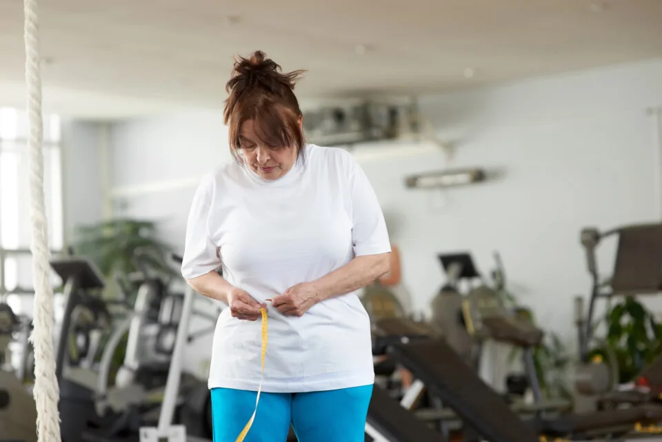 A woman in a gym measures her waist with a tape measure, tracking her weight loss progress amid various exercise equipment, mindful of how menopause can affect fitness goals.