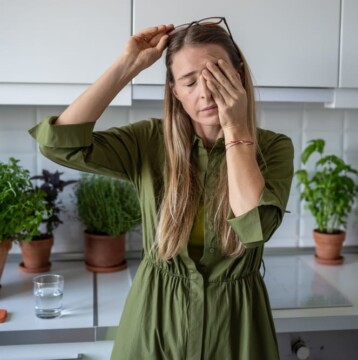 A woman in a green dress holds her glasses and rubs her eyes in the kitchen, signs of perimenopause tiredness. Various potted plants fill the background, while a glass of water sits on the counter.