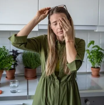 A woman in a green dress holds her glasses and rubs her eyes in the kitchen, signs of perimenopause tiredness. Various potted plants fill the background, while a glass of water sits on the counter.