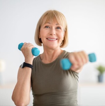 A person with short blonde hair exercises with blue dumbbells, wearing a grayish-green shirt and a smartwatch, against a soft-focus neutral background.