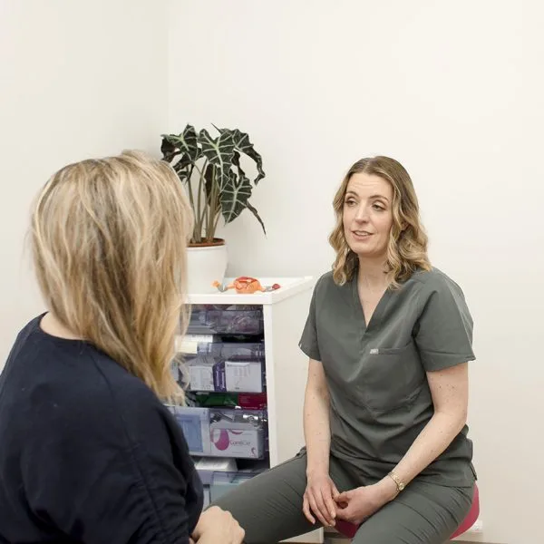 Two women engage in a Private Women's Health Consultation in a medical office; one in scrubs speaks while the other listens. A plant and medical supplies are visible in the background.