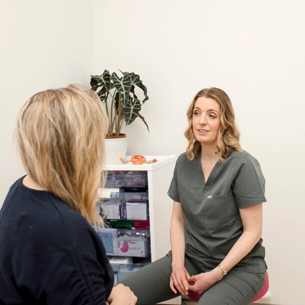 Two women engage in a Private Women's Health Consultation in a medical office; one in scrubs speaks while the other listens. A plant and medical supplies are visible in the background.
