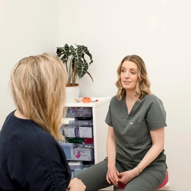 Two women engage in a Private Women's Health Consultation in a medical office; one in scrubs speaks while the other listens. A plant and medical supplies are visible in the background.