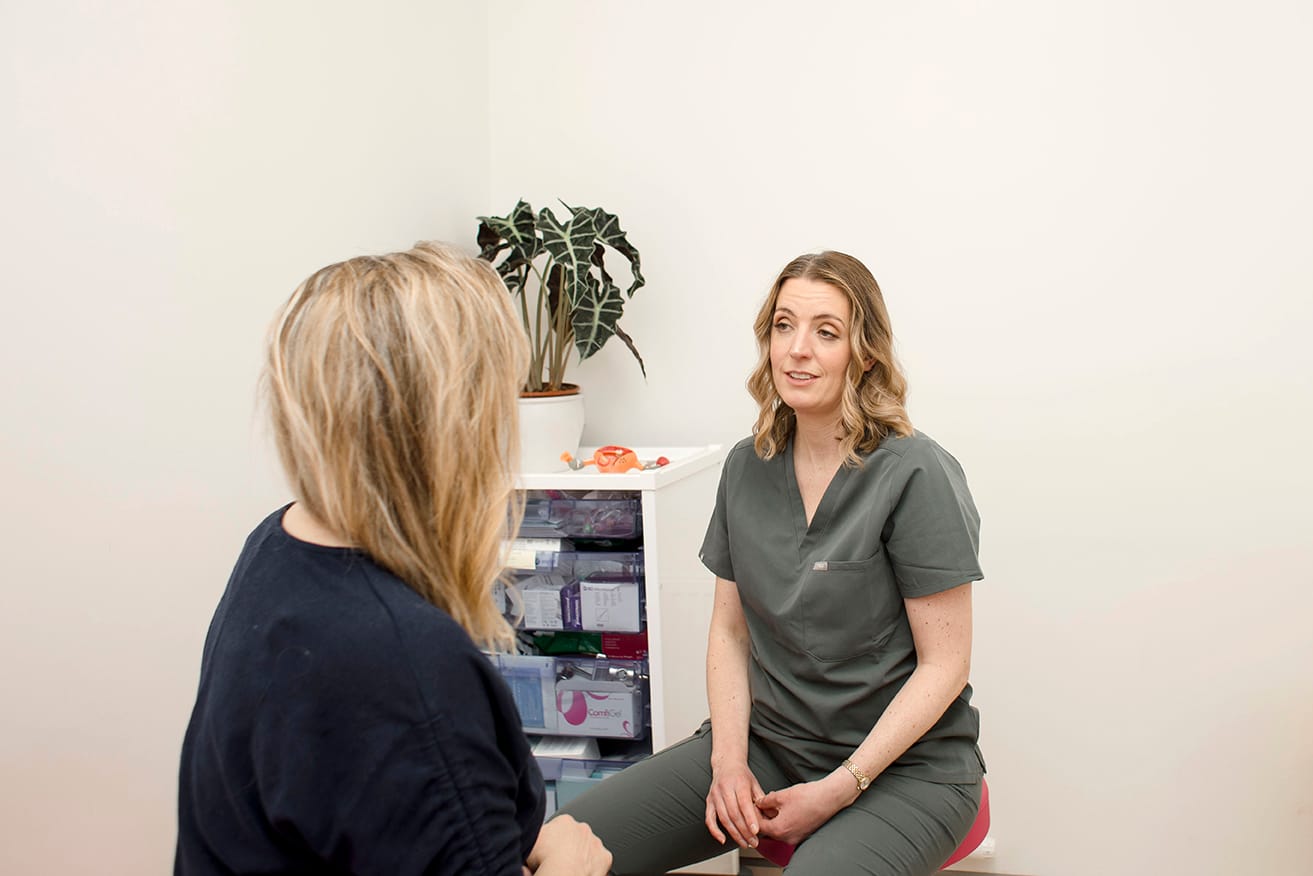 Two women engage in a Private Women's Health Consultation in a medical office; one in scrubs speaks while the other listens. A plant and medical supplies are visible in the background.