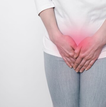 A person in a white shirt and gray pants stands against a white background, clutching their lower abdomen. The red overlay suggests discomfort, possibly linked to menopause or issues like vaginal atrophy.