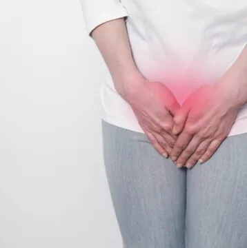 A person in a white shirt and gray pants stands against a white background, clutching their lower abdomen. The red overlay suggests discomfort, possibly linked to menopause or issues like vaginal atrophy.