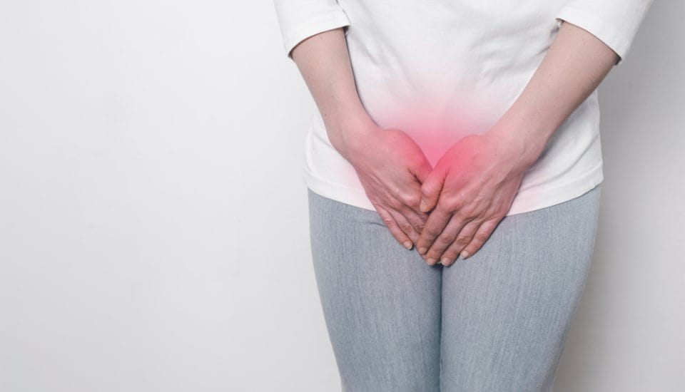 A person in a white shirt and gray pants stands against a white background, clutching their lower abdomen. The red overlay suggests discomfort, possibly linked to menopause or issues like vaginal atrophy.