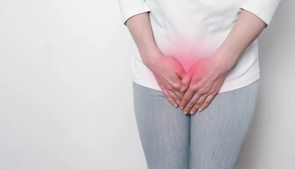 A person in a white shirt and gray pants stands against a white background, clutching their lower abdomen. The red overlay suggests discomfort, possibly linked to menopause or issues like vaginal atrophy.