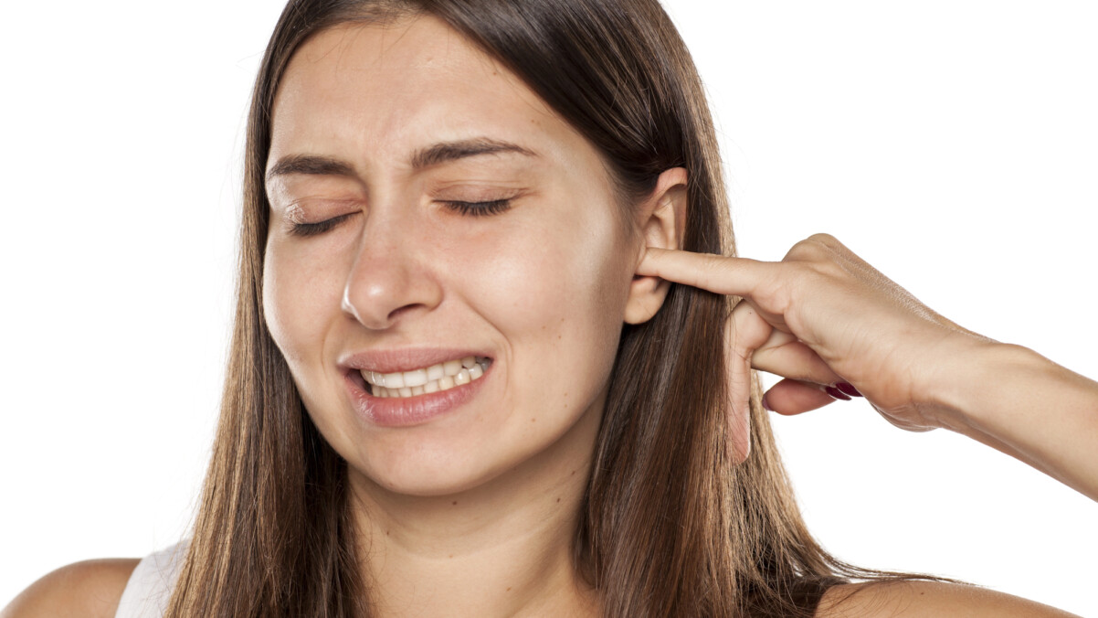 A woman with long brown hair closes her eyes and grimaces while inserting a finger into her itchy ear against a white background.