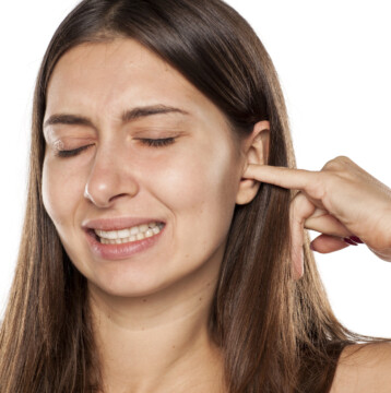 A woman with long brown hair closes her eyes and grimaces while inserting a finger into her itchy ear against a white background.