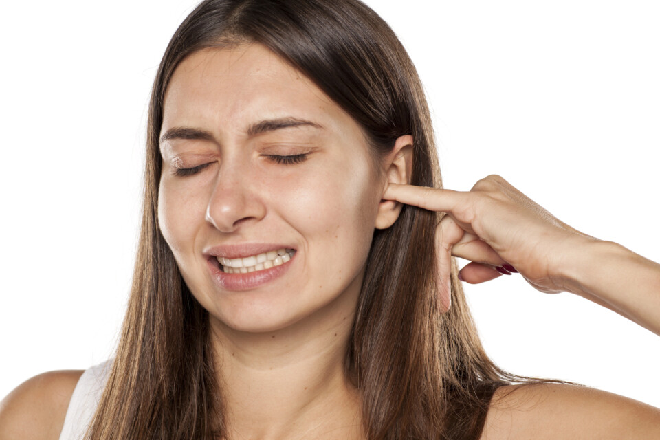 A woman with long brown hair closes her eyes and grimaces while inserting a finger into her itchy ear against a white background.
