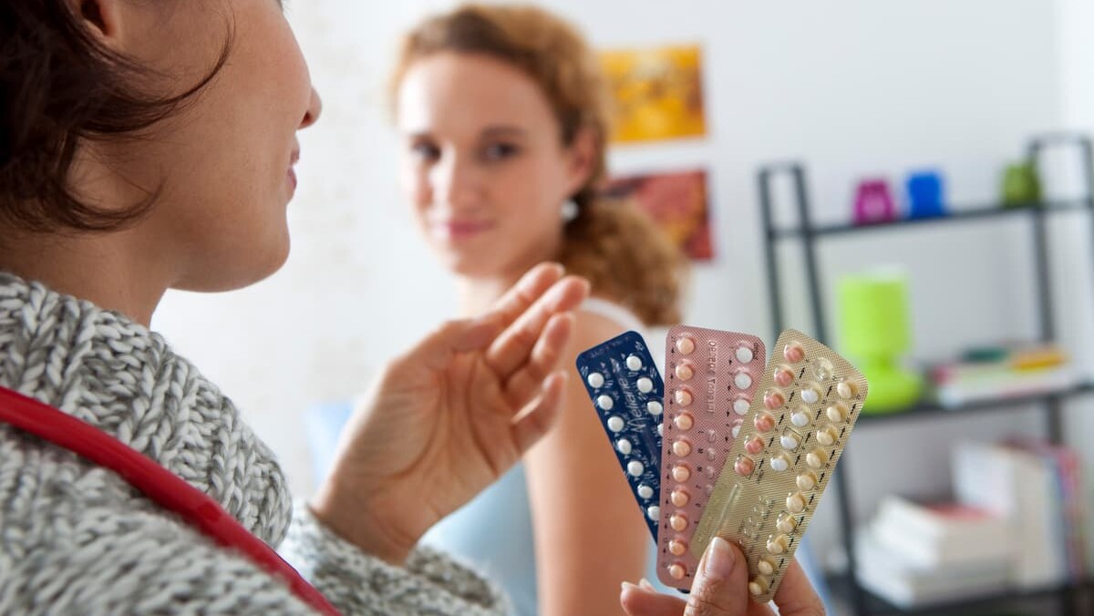 A healthcare professional shows different packs of birth control pills, including the Progestogen-Only Pill, to a patient in a medical office and explains possible side effects such as desogestrel bleeding.