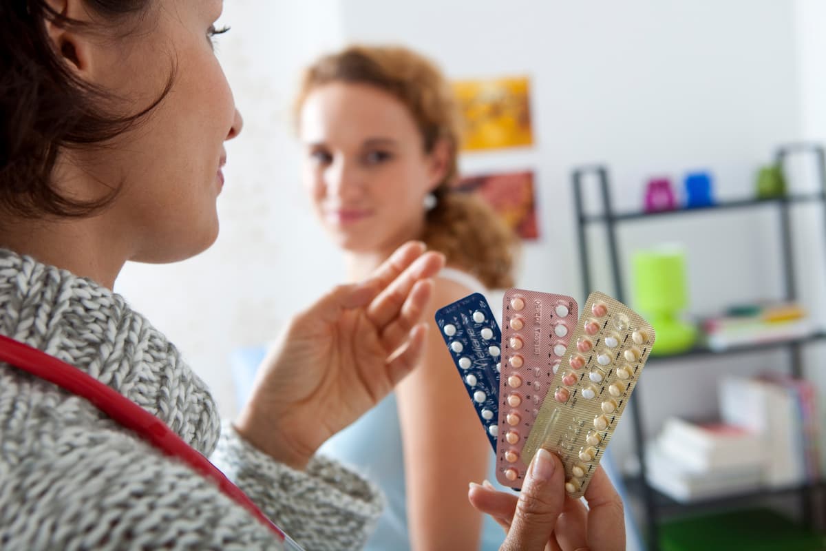 A healthcare professional shows different packs of birth control pills, including the Progestogen-Only Pill, to a patient in a medical office and explains possible side effects such as desogestrel bleeding.