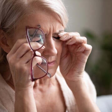 Older woman with light hair holds her glasses in one hand and rubs her eyes with the other, appearing tired or experiencing eye discomfort—possibly a sign of dry eyes often linked to menopause.