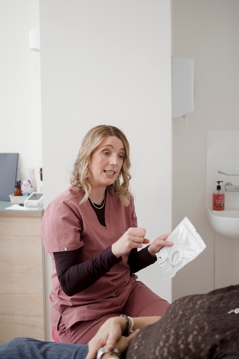 A healthcare worker in pink scrubs explains a vaginal pessaries package to a patient in a clinical setting.
